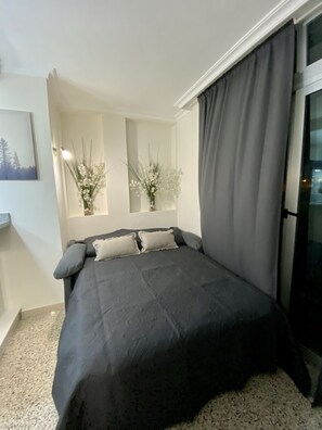 A bedroom with a large bed, two bedside tables, a window with curtains, and a wall-mounted light fixture.