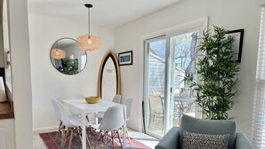A dining area with a white table, chairs, and a red patterned rug. There is a grey armchair with a patterned pillow, a tall green plant, and a large mirror on the wall. - UPDATED IN 2022  @Oceanfront - 3 Blocks 2Beach! 2 Suites 2.5 bath - great deck! (Virginia Beach)