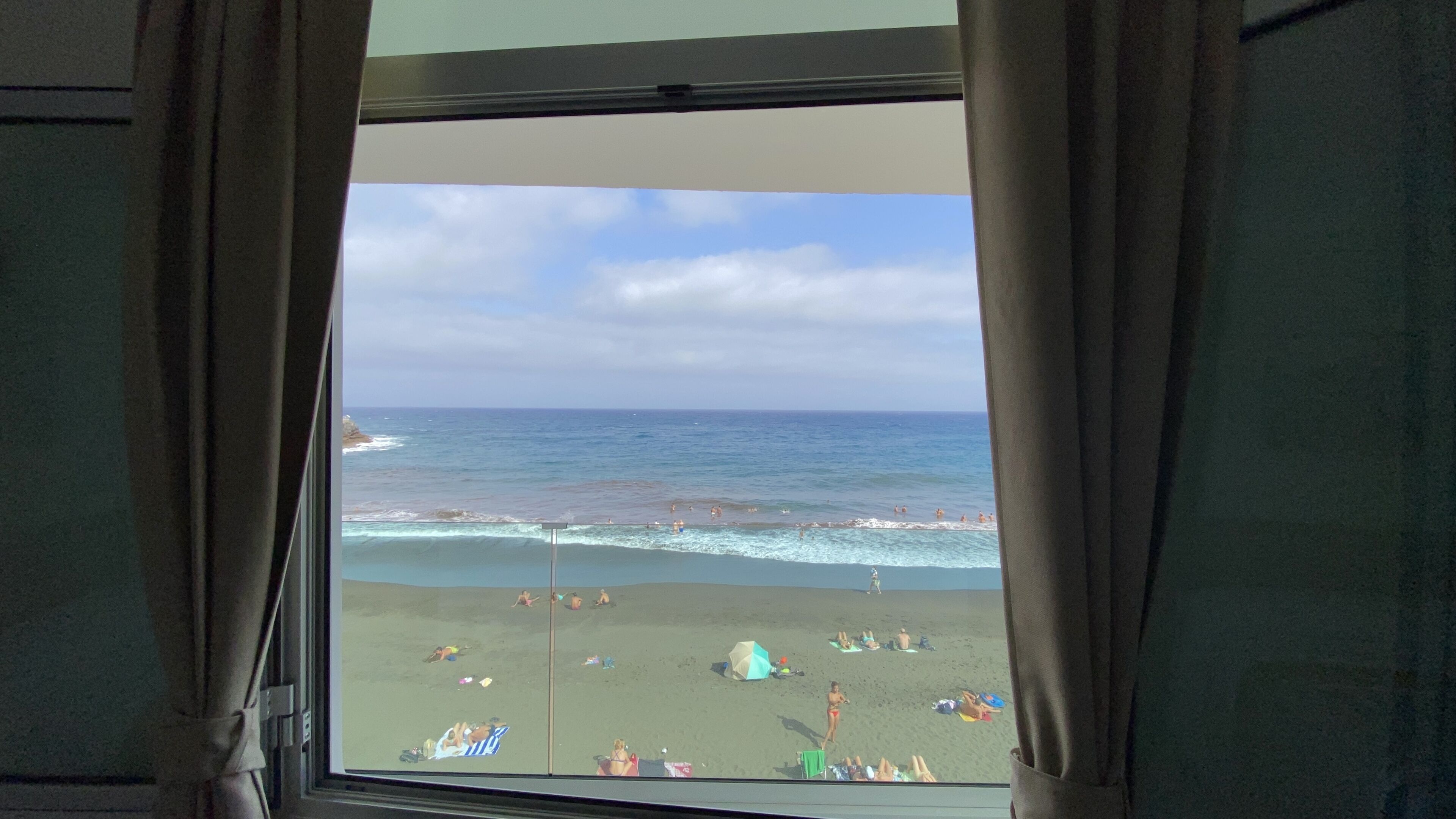 A beach view through a window with curtains, featuring the ocean, beachgoers, and a clear sky.