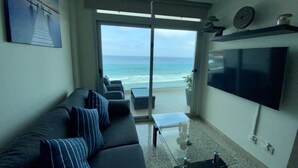 Uma sala de estar com um sofá, uma mesa de centro e uma televisão. Há uma porta de vidro deslizante que dá para uma varanda com vista para o oceano.