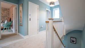 A hallway with a carpeted floor, a white door, and a staircase with a wooden handrail. - 6 Bedroom New Build Detached House in Bicester (Bicester)