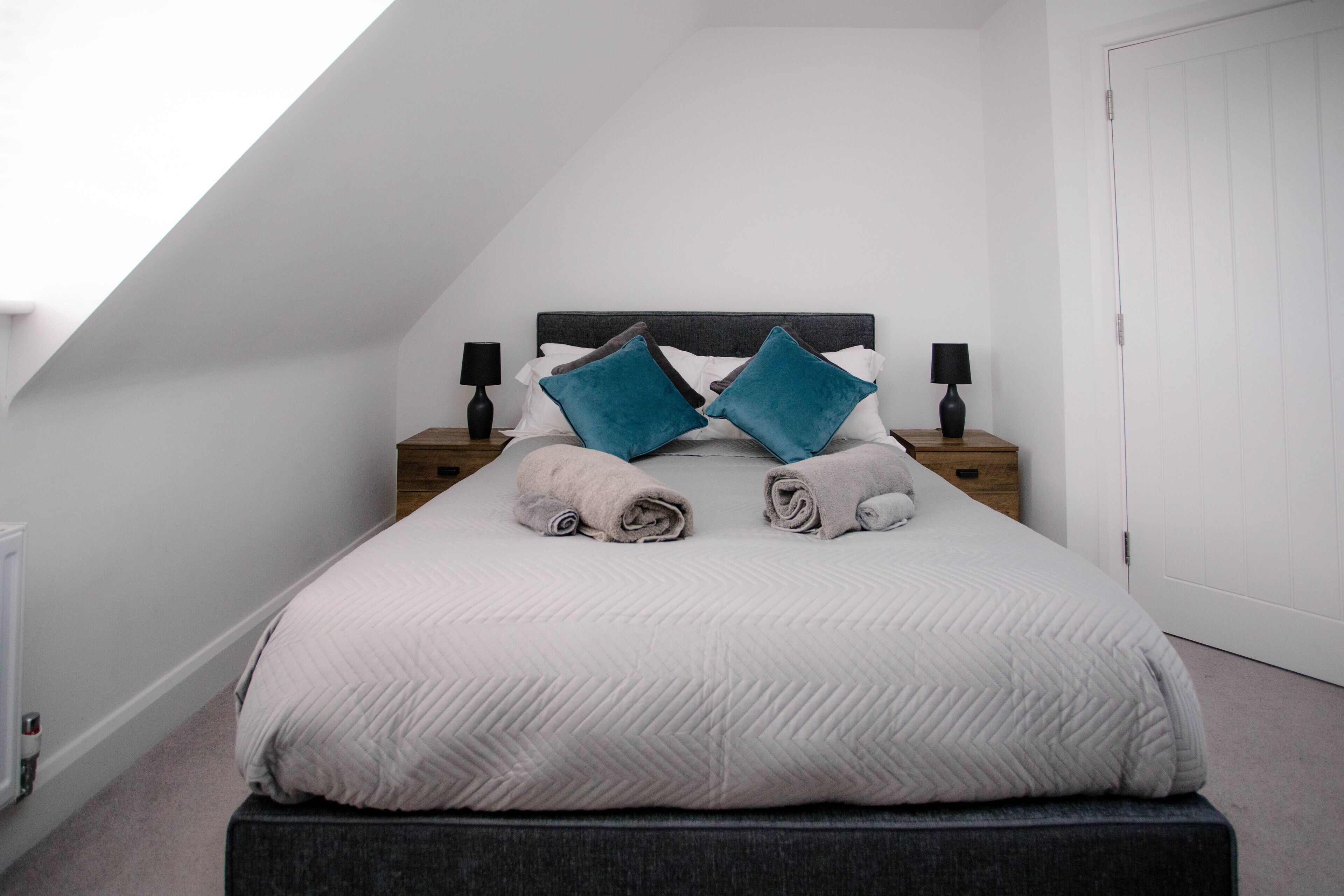 A neatly made bed with a grey headboard, two bedside tables with lamps, and a white door in a room with white walls and a sloped ceiling.