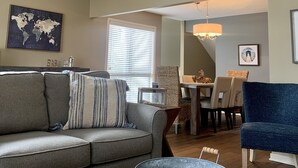 A spacious living room with a high ceiling, a ceiling fan, a world map artwork, a dining area with a chandelier, and a large window with blinds.