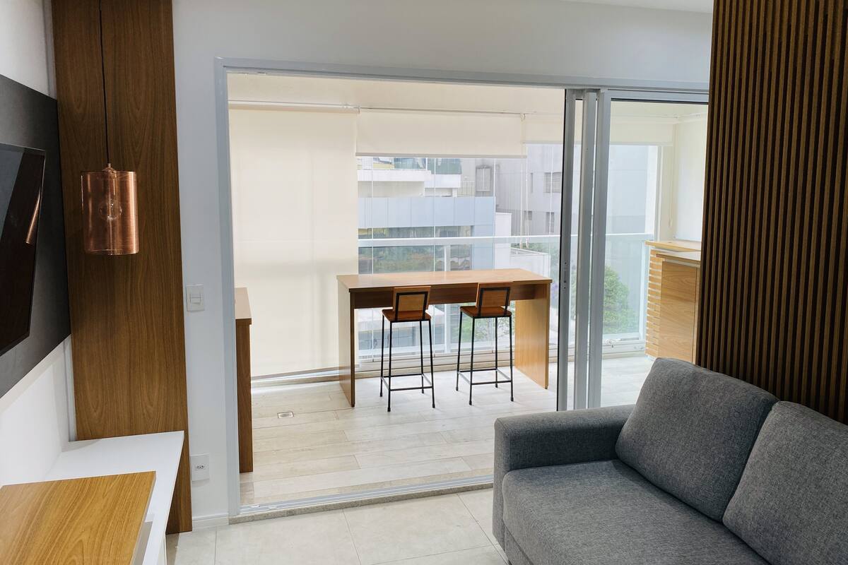 Una sala de estar moderna con un sofá gris, una mesa de comedor de madera con dos sillas y un gran ventanal corredera que da a un balcón con vistas a otros edificios.