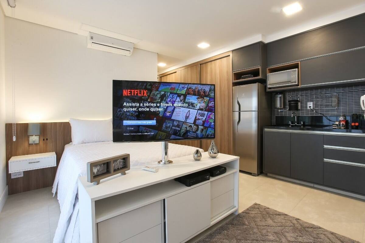 Una habitación de hotel moderna con una televisión de pantalla plana grande, una cama, una mesita de noche, un armario y una kitchenette.