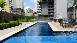 Una piscina con suelo de baldosas, rodeada por un muro bajo y follaje. Hay sillones de descanso y sombrillas en el lado derecho.