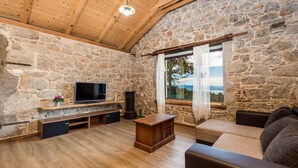 A rustic stone-walled room with a wooden ceiling, a television on a shelf, a sofa, and a window with a view. - Cozy home in Veprinac (Veprinac)