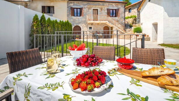 An outdoor dining area with a table set for two, featuring a white tablecloth with olive branch patterns, a plate of strawberries, a bowl of cherries, a glass of white wine, and a croissant. - Cozy home in Veprinac (Veprinac)
