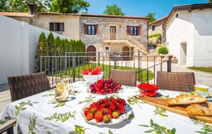 An outdoor dining area with a table set for two, featuring a white tablecloth with olive branch patterns, a plate of strawberries, a bowl of cherries, a glass of white wine, and a croissant. - Cozy home in Veprinac (Veprinac)