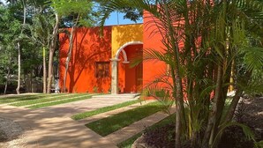 Un bâtiment à l’extérieur rouge, un chemin pavé et un jardin bien entretenu avec des palmiers et des arbustes.
