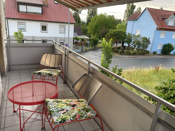 Un balcon avec deux chaises et une table, donnant sur une rue résidentielle bordée de maisons et d’espaces verts.