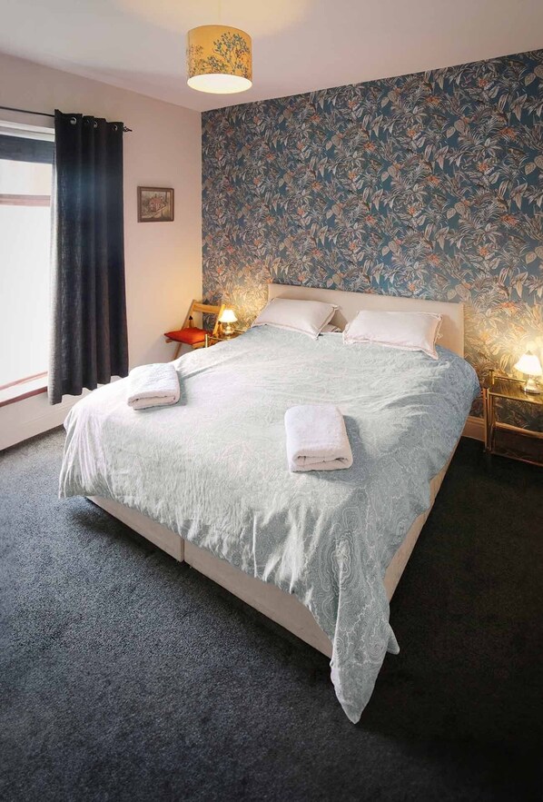 A bedroom with a bed, two bedside lamps, a window with curtains, and a floral wallpaper.
