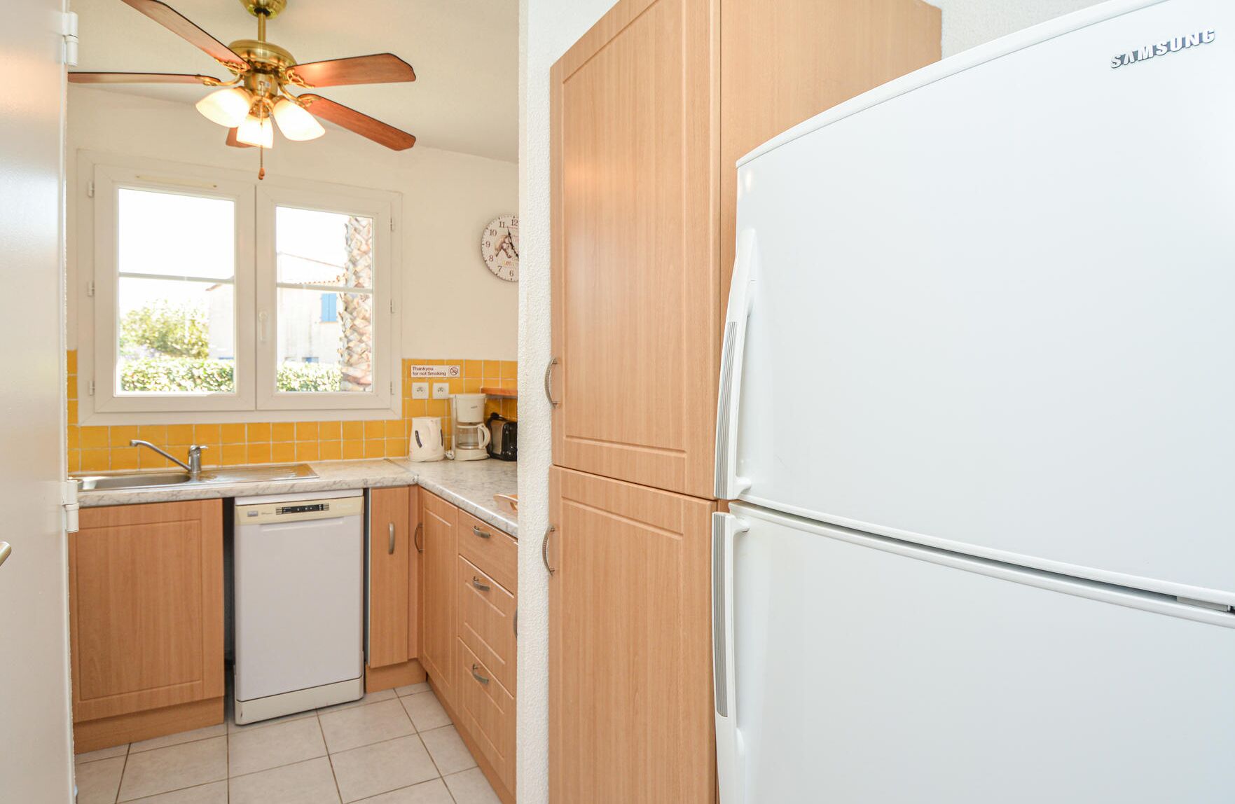 A kitchen with wooden cabinets, a white refrigerator, a dishwasher, a ceiling fan, and a window with a view of greenery.