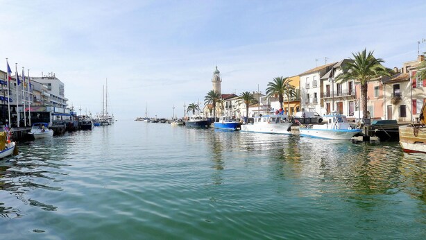 Un puerto deportivo con barcos amarrados a lo largo de la costa, edificios con balcones y una torre destacada al fondo.