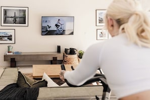 Un salon avec un téléviseur diffusant une vidéo de cyclisme, un canapé, une table basse et un mur orné de tableaux encadrés.
