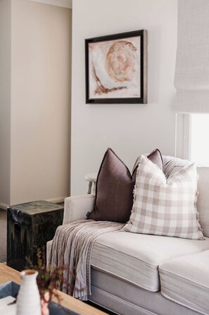 A modern living room with a sofa, cushions, a framed artwork, and a window with blinds.