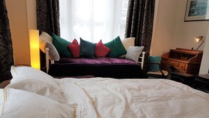 Ein ordentlich bezogenes Bett mit weißer Bettwäsche und goldener Zierleiste, ein Sofa mit bunten Kissen, eine Lampe und ein Holznachttisch.