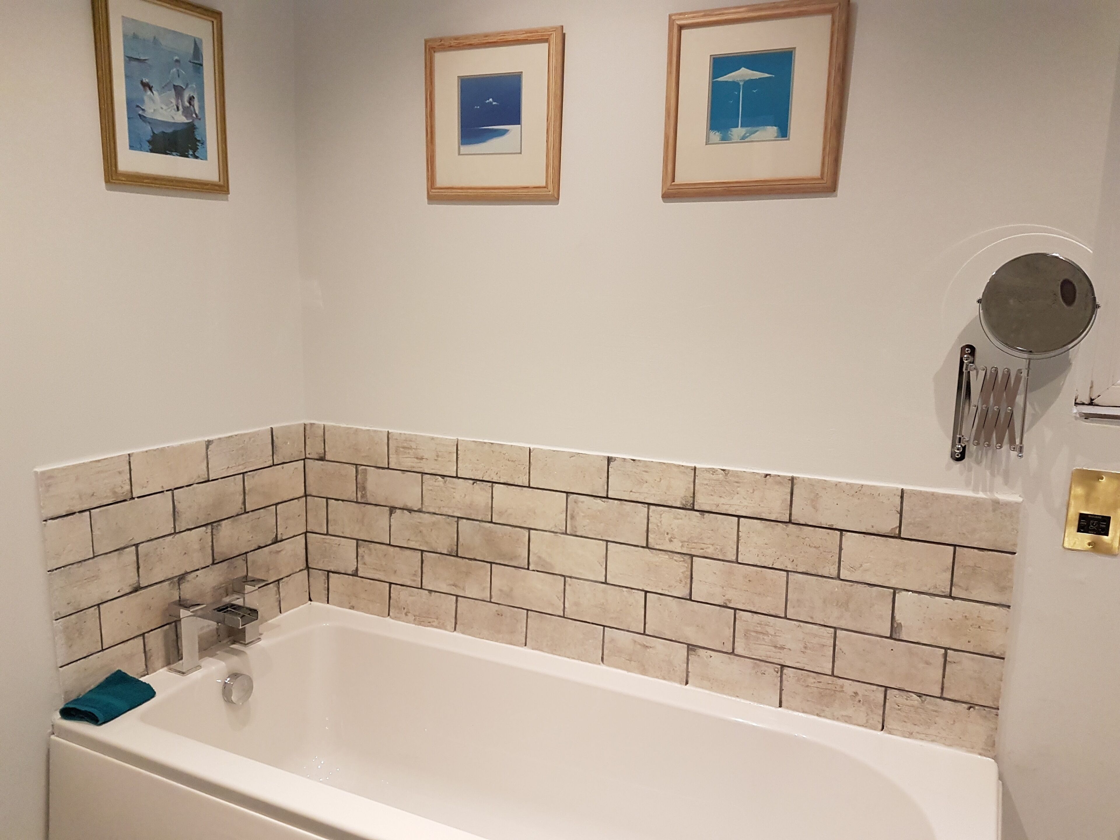 Ett badrum med ett vitt badkar, beige tegelstensplattor på väggen och tre inramade tavlor på väggen.