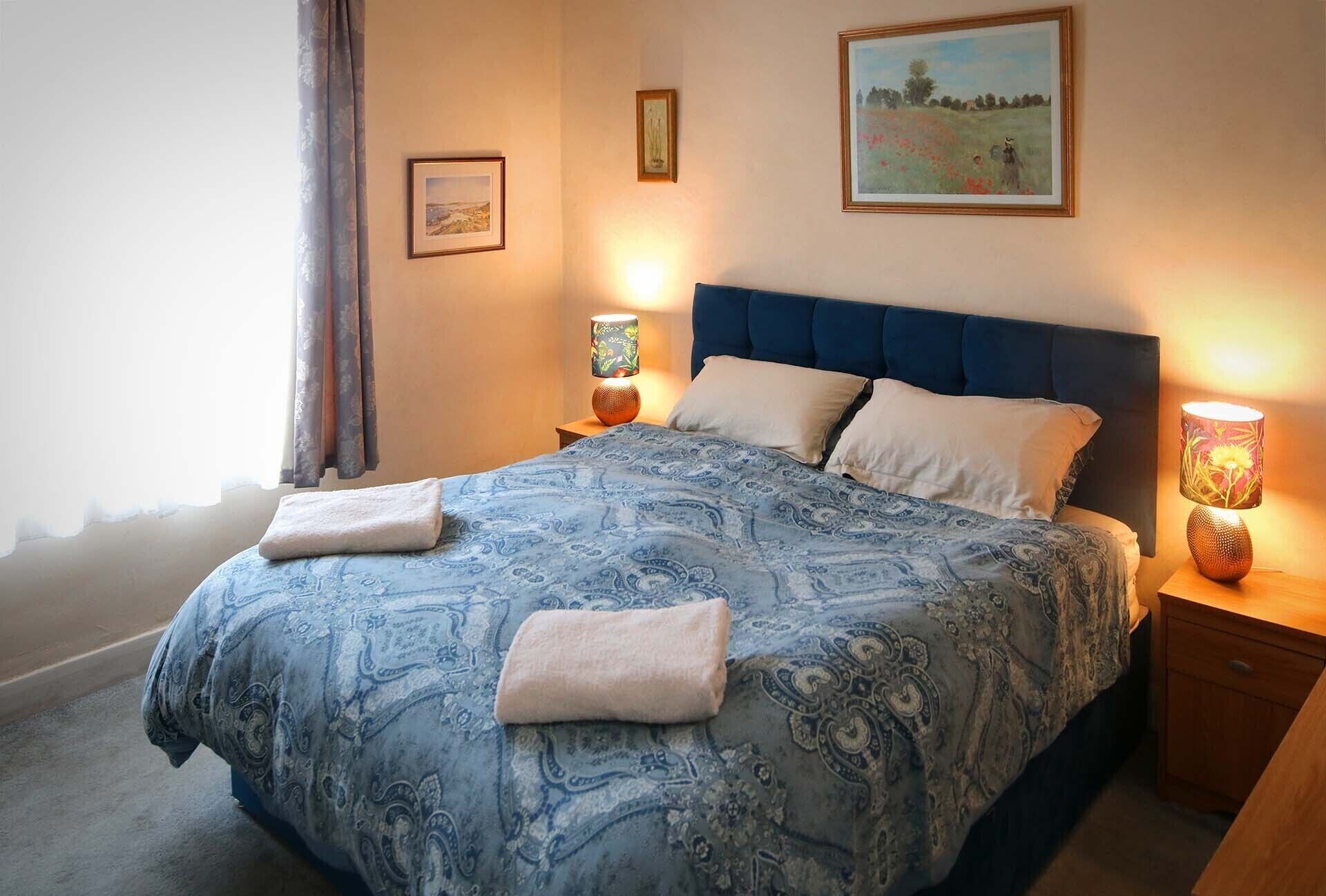 Un letto rifatto con cura, coperta azzurra a motivi decorativi e due cuscini, affiancato da due abat-jour con decori floreali.