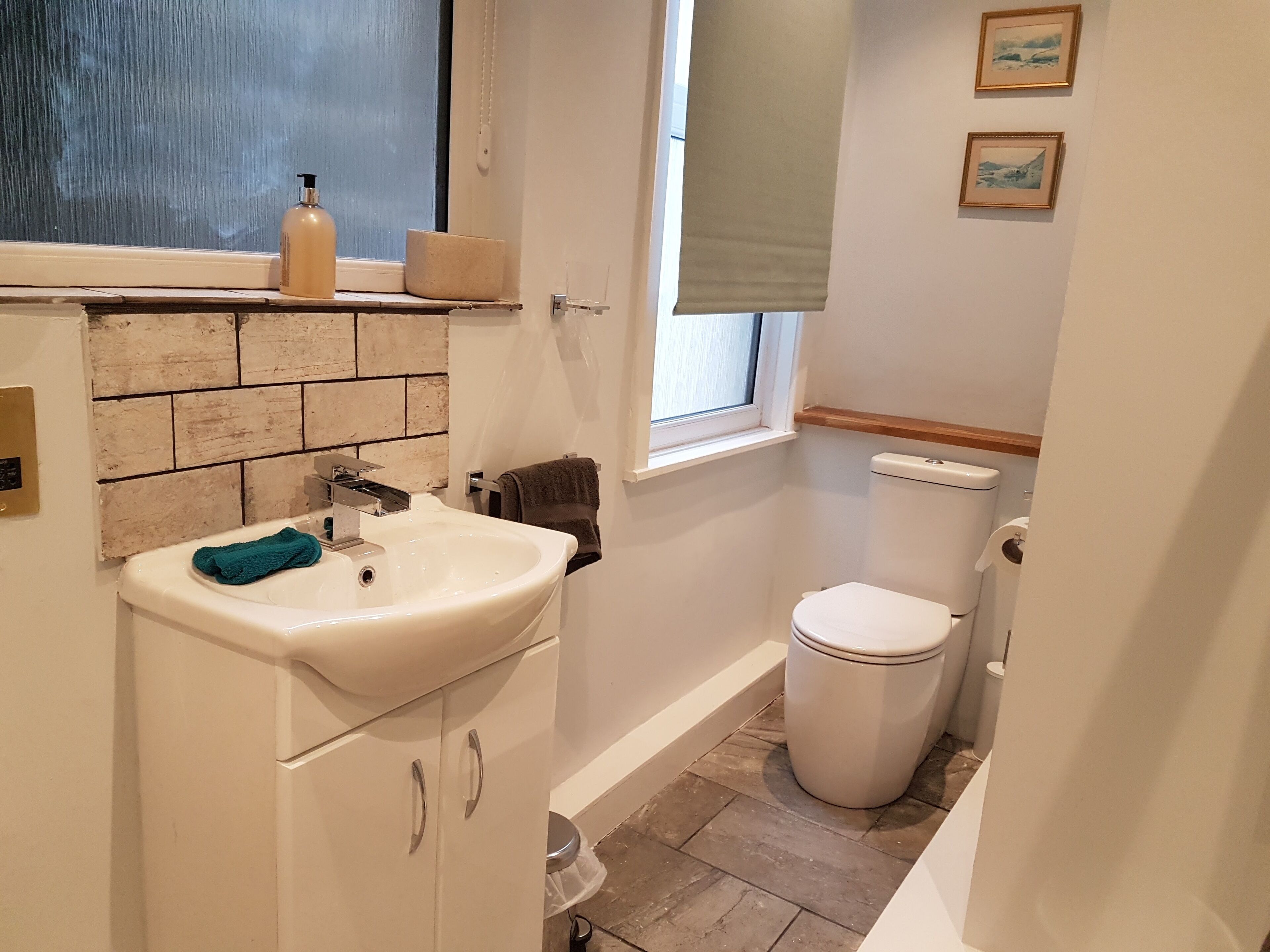 A bathroom with a white sink, a toilet, a window with a blind, and a small shelf with toiletries.