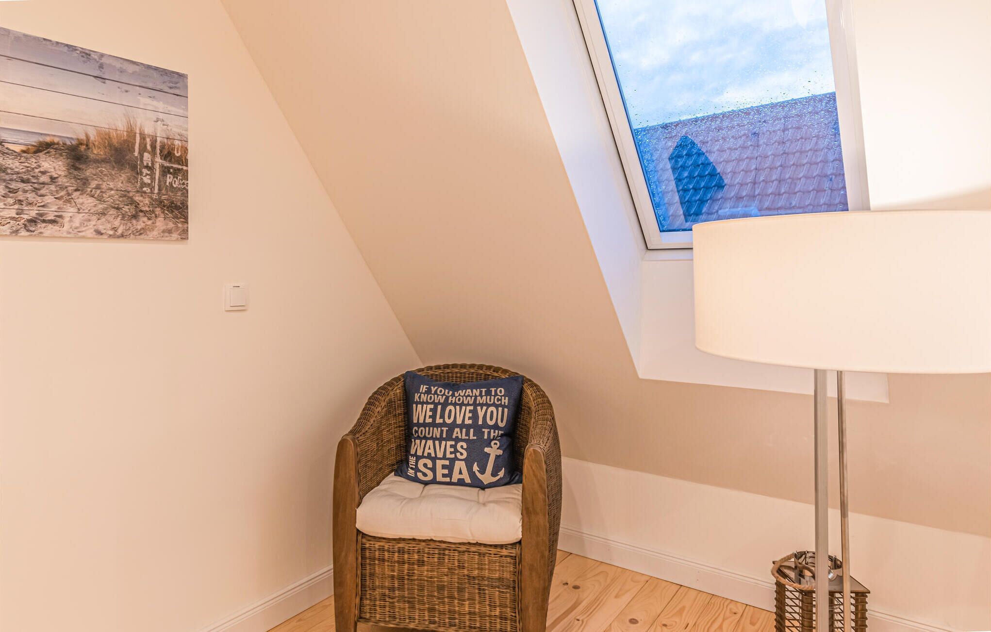 Una habitación de esquina con techo inclinado, una silla de mimbre con cojín, una mesita y un cuadro en la pared.
