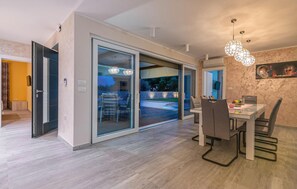 Un espace repas moderne doté d’une porte coulissante en verre donnant sur un espace extérieur, d’une table à manger avec des chaises et d’un lustre.