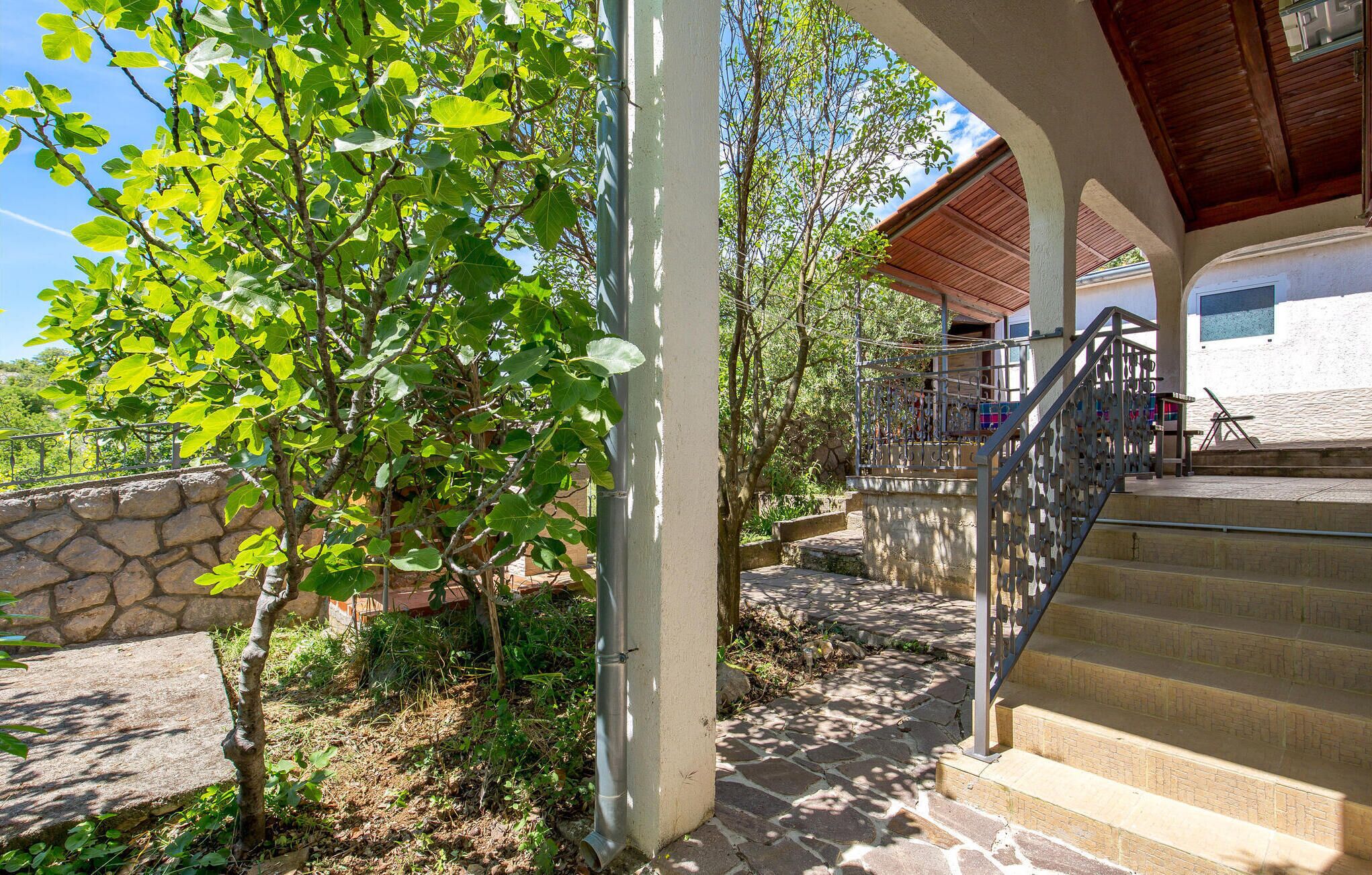 A stone staircase leading to a wooden deck with a metal railing, surrounded by greenery and a stone wall.
