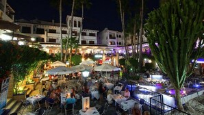 An outdoor dining area with tables and umbrellas, surrounded by palm trees and buildings. - 1 Bed 1 Bath Luxurious Apt Villamartin Plaza, Golf, Fun & Sun (Orihuela Costa)
