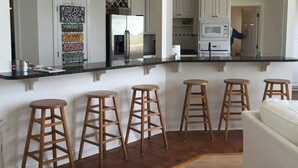 Eine moderne Küche mit weißen Schränken, einem Edelstahlkühlschrank und einem eingebauten Backofen. Im Sitzbereich befinden sich vier Holzstühle und ein weißes Sofa.