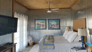 Ein Schlafzimmer mit Holzboden, zwei gerahmten Bildern, einem Bett mit weißer Bettwäsche, einem Fernseher auf einem Holzständer und einem Fenster mit Vorhängen.