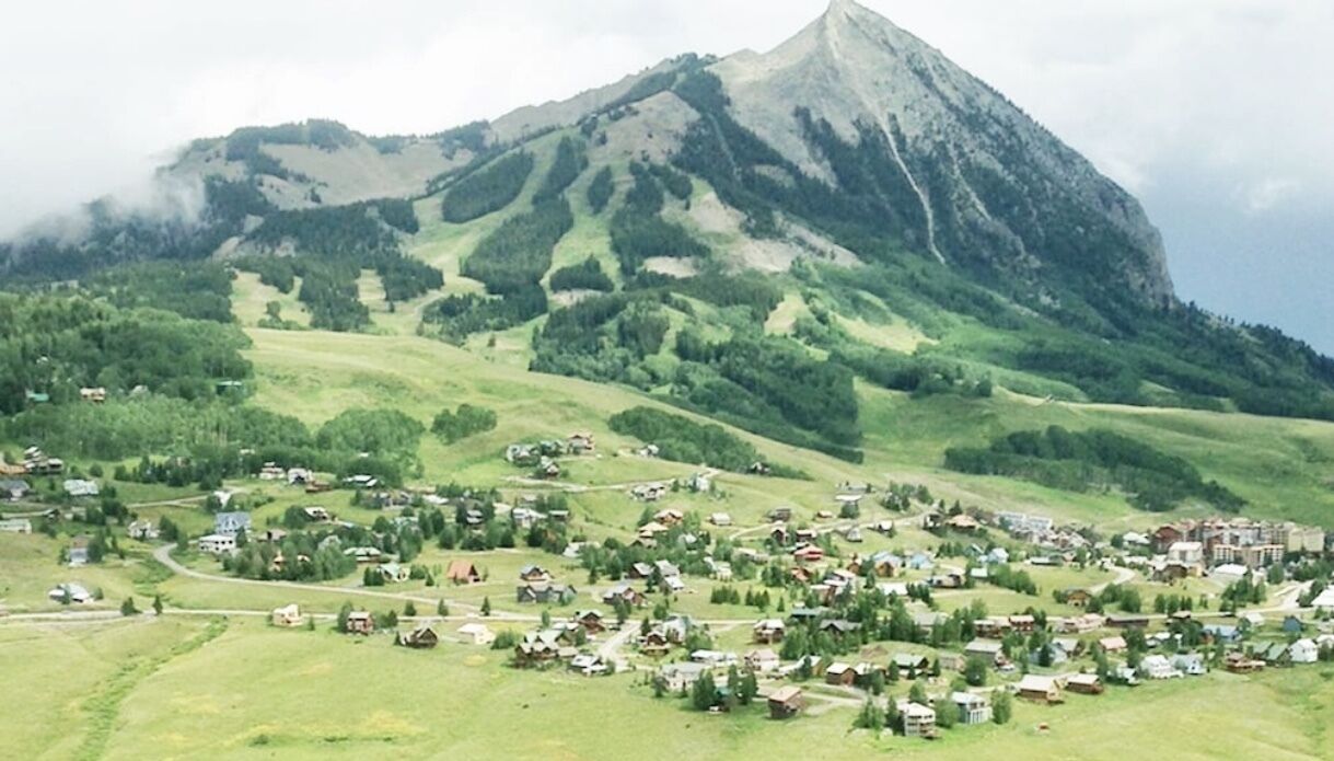 A rural village with houses spread across a green valley, surrounded by hills and a prominent mountain in the background.