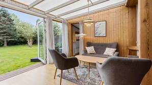 A modern living room with a glass door leading to a garden, wooden walls, a grey sofa, and a table with chairs. - 3 bedroom pet friendly home in Hundested (Hundested)