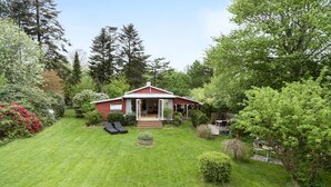 A red cabin with a covered porch, surrounded by lush greenery and trees. - 3 bedroom pet friendly home in Hundested (Hundested)