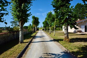 Une étroite route pavée, bordée d’arbres et d’un mur de pierres d’un côté, mène à un bâtiment dont le toit est recouvert de tuiles.