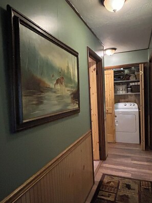 A hallway with a framed painting of deer by a river, a wooden door leading to a laundry area, and a radiator on the wall.