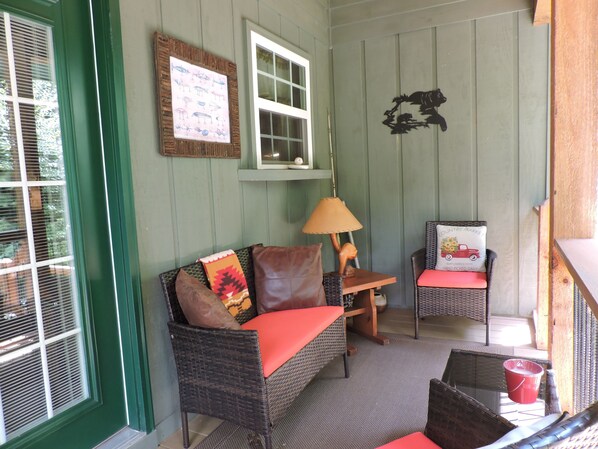 Une pièce chaleureuse avec un canapé en osier, une petite table et une chaise. On y trouve une fenêtre avec des stores, une lampe et un élément décoratif mural.