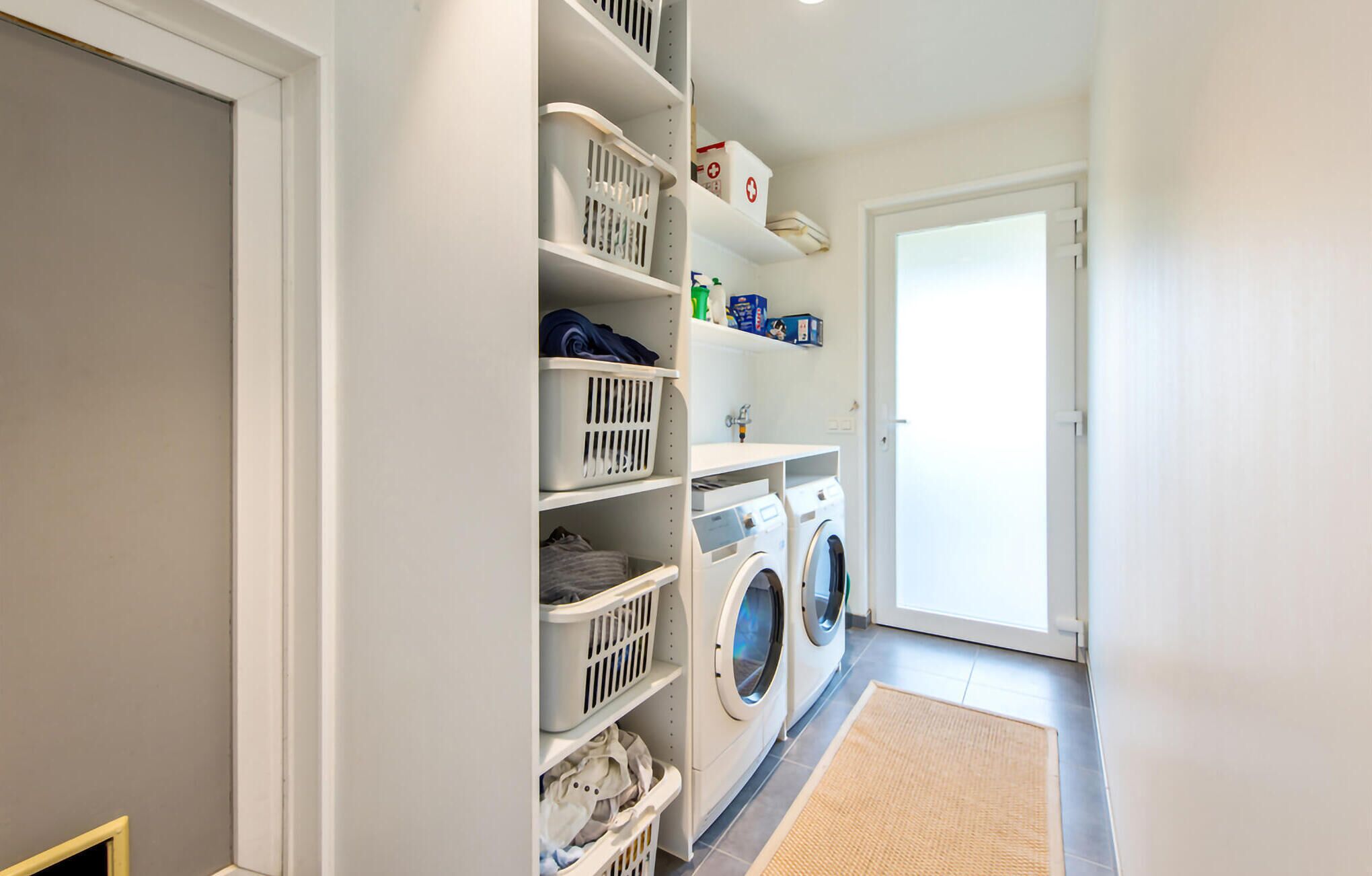 Une buanderie équipée d’un lave-linge et d’un sèche-linge, de plusieurs étagères où sont rangés des paniers à linge, et d’une porte donnant sur une autre pièce.