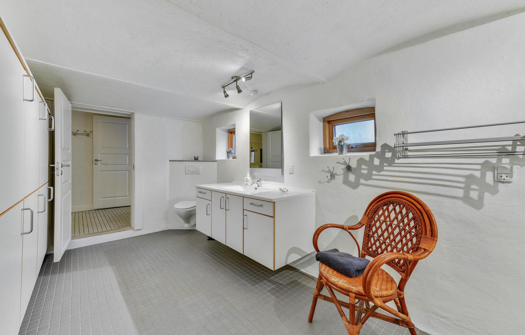 A bathroom with a white sink, mirror, and toilet. There is a wicker chair with a towel on it.