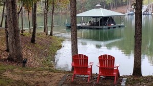 A tranquil lakeside scene with a docked boat, two red chairs, and a gazebo overlooking the water.
