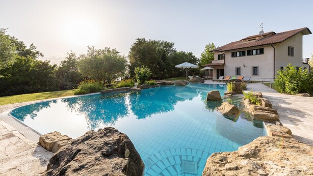 Uma piscina com água azul cristalina, rodeada por pedras e vegetação, e um edifício com telhado de azulejos ao fundo.