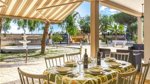 Un patio exterior con una mesa lista para comer, rodeado de sillas y una zona cubierta con un toldo a rayas.