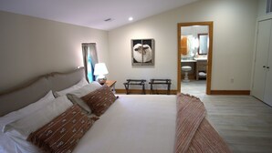 Una camera da letto con un letto grande, un comodino con una lampada, un quadro appeso al muro e un bagno visibile attraverso una porta aperta.