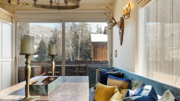 A cozy living room with a wooden table, a sofa with colorful cushions, a chandelier, and a deer head mounted on the wall.