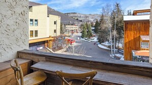 A balcony with wooden chairs and a view of a town with buildings, a red vehicle, and snow-covered ground.