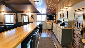 Un restaurant doté d’un long comptoir en bois, de tabourets de bar et d’un espace cuisine avec des placards et un réfrigérateur.