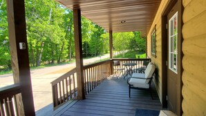 Une véranda couverte avec une balustrade en bois, une table avec deux chaises et une vue sur une route et des arbres.