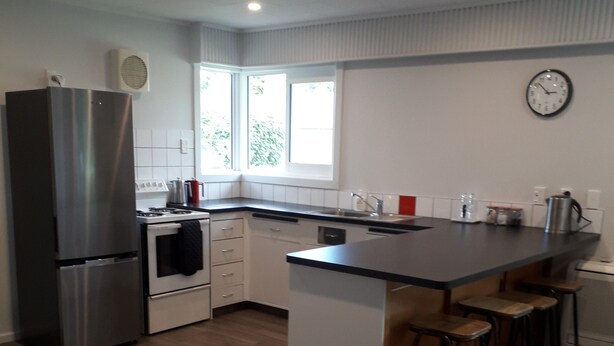 A modern kitchen with a stainless steel refrigerator, a stove, and a sink. There are wooden stools at the island and a clock on the wall. - Fairlie Family Home (Fairlie)