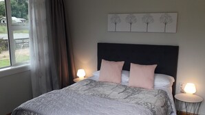 A bedroom with a large bed, a headboard, two bedside lamps, a window with curtains, and a wall-mounted artwork of trees. - Fairlie Family Home (Fairlie)