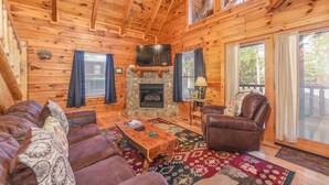 A cozy cabin interior with a stone fireplace, wooden walls, a ceiling fan, and a large window. - Amazing Log Chalet, Scenic, Mountain View’s, Quiet (Sevierville)