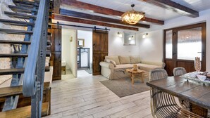 Een rustiek interieur met zichtbare houten balken, een houten trap, een bank, een salontafel en een eettafel met stoelen.